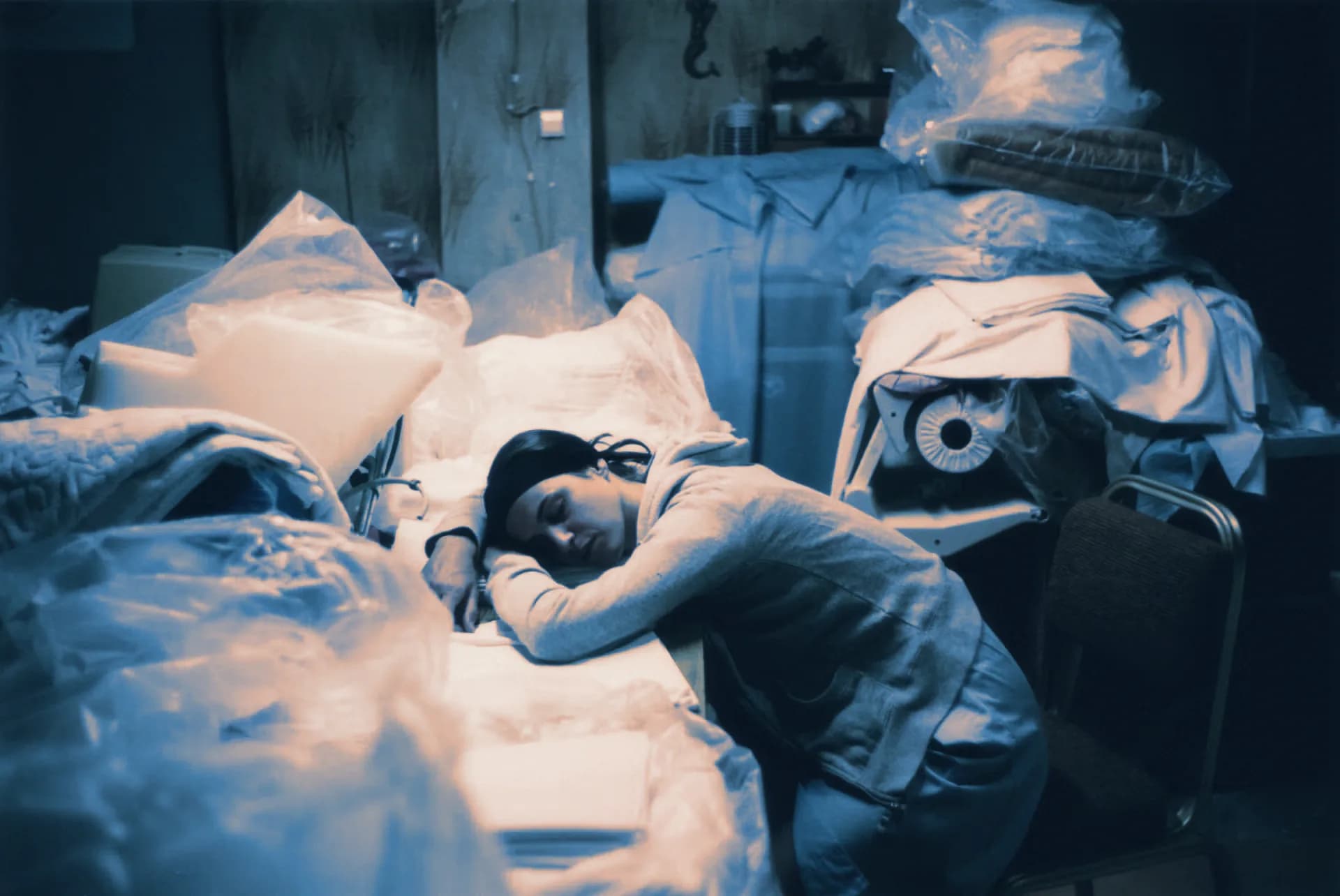 A woman asleep at a table piled with plastic-wrapped linen, in blue light